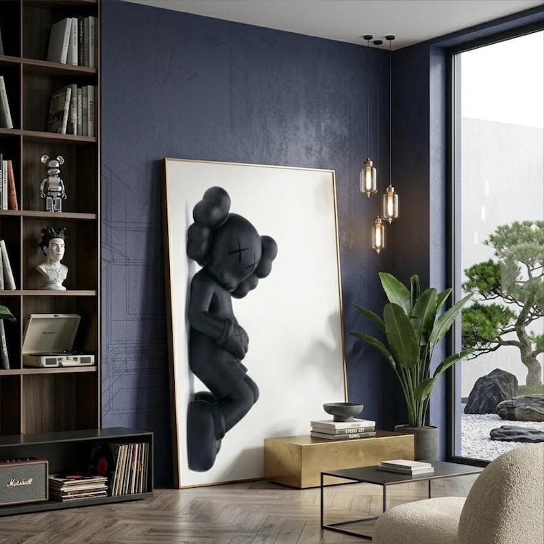 May include: A large framed artwork featuring a black sculpture of a figure with crossed-out eyes and a rounded head. The artwork is mounted on a white background with a gold frame. The room has a modern design with a dark blue wall and a wooden bookshelf.