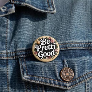 Pode incluir: Um botão redondo com as palavras "Be Pretti Good" em escrita branca sobre um fundo preto, rodeado por detalhes florais. O botão é bege e está preso a uma jaqueta jeans.