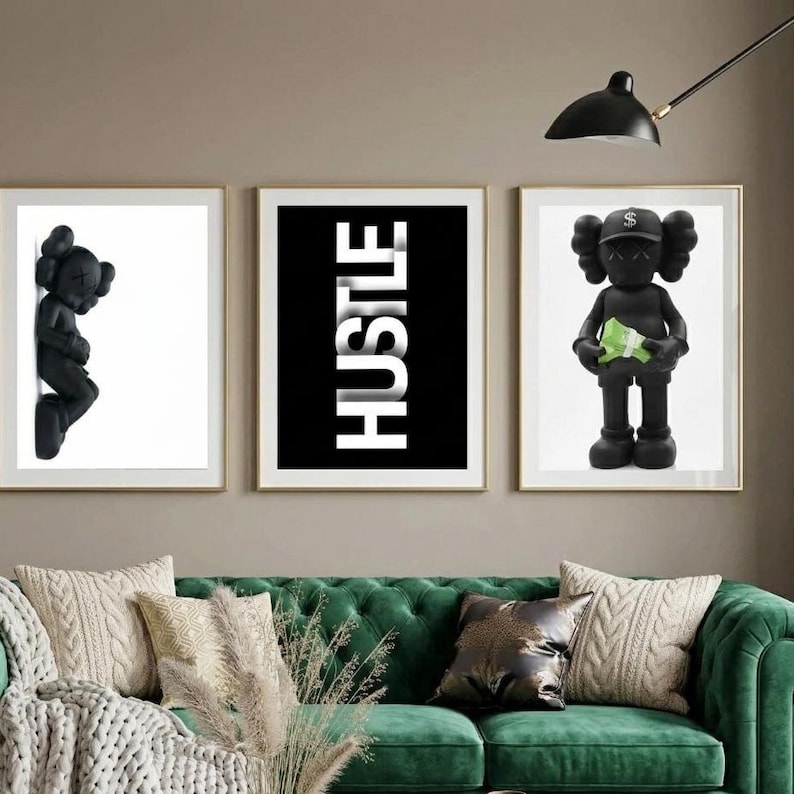 May include: Three framed art prints. The left print features a black figurine on a white background. The center print has the word "HUSTLE" in white on a black background. The right print shows a black figurine holding green money. The prints are above a green sofa.