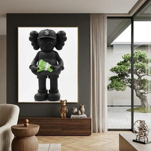 May include: A large framed artwork featuring a black figure with crossed-out eyes and a dollar sign cap, holding a bundle of green money. The figure is set against a white background. The artwork is displayed in a modern living room setting.