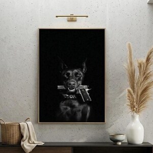 May include: Black and white framed print featuring a Doberman Pinscher holding a handgun in its mouth. The dog is in the center of the image, set against a dark background. The print is displayed on a light gray wall.