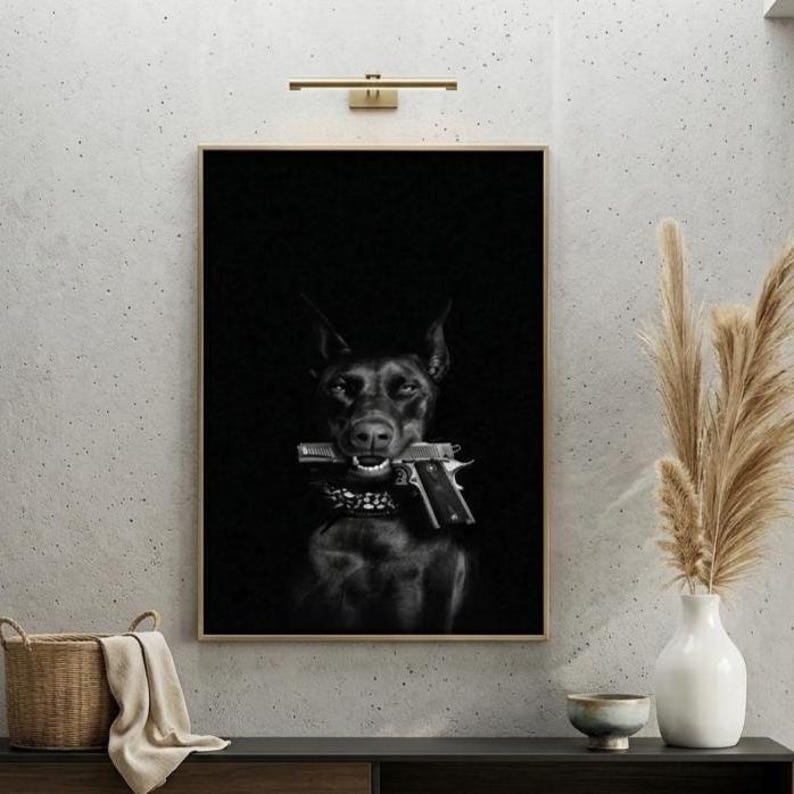 May include: Black and white framed print featuring a Doberman Pinscher holding a handgun in its mouth. The dog is in the center of the image, set against a dark background. The print is displayed on a light gray wall.