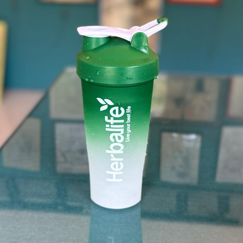 Puede incluir: Una botella mezcladora Herbalife verde y blanca con tapa y tapa abatible blancas. La botella tiene un dise&ntilde;o degradado, que pasa de verde a blanco. El logotipo de Herbalife y el texto "Live your best life" est&aacute;n impresos en la parte delantera.