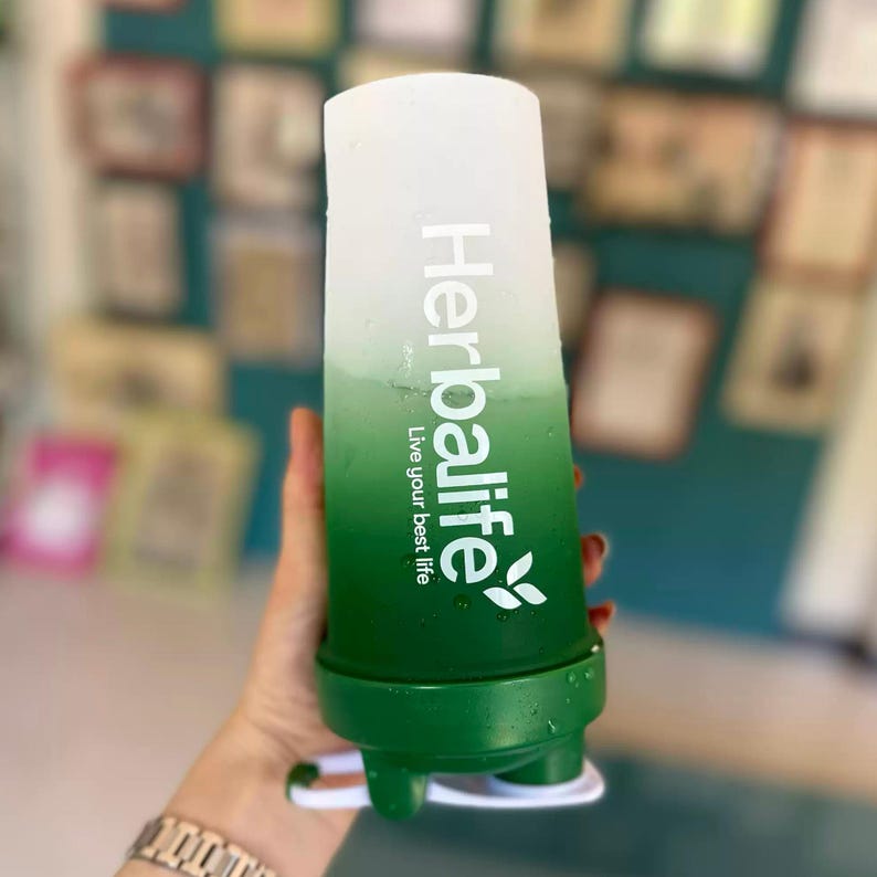 Puede incluir: Una botella mezcladora degradada blanca y verde con el texto "Herbalife" y "Live your best life". La botella tiene una base verde y un asa blanca.