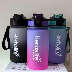 May include: Three water bottles with a gradient design. The bottles are black, pink, and purple with the Herbalife logo and the text "Live your best life".