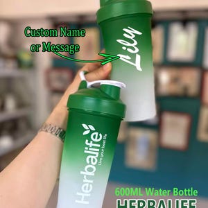 Puede incluir: Dos botellas de agua con degradado verde y blanco. Una botella tiene el nombre "Lily" en escritura blanca. La otra botella tiene el logotipo y el texto "Herbalife". Las botellas est&aacute;n etiquetadas como "600ML Water Bottle" y "Custom Name or Message".