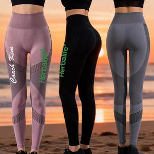 May include: Three pairs of athletic leggings in mauve, black, and grey. The mauve leggings have the text "Coach Kim" and "Herbalife" printed on them. The black leggings have "Herbalife" printed on them. The grey leggings have a contrasting grey panel.