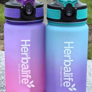 May include: Two ombre water bottles with black lids and purple and blue color gradients. The bottles have the Herbalife logo and the text "Live your best life" printed on them.