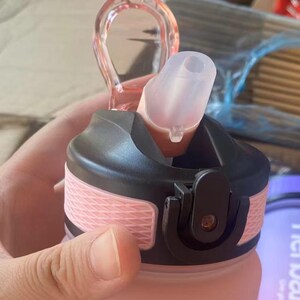 May include: Pink water bottle with a black lid and a clear silicone straw. The bottle has a textured design and a black carrying loop.