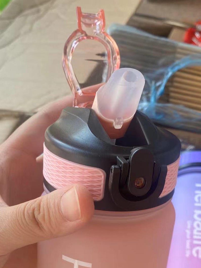 May include: Pink water bottle with a black lid and a clear silicone straw. The bottle has a textured design and a black carrying loop.