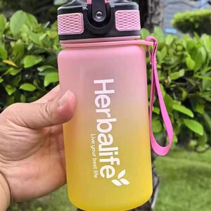 May include: A pink and yellow water bottle with a black lid and a pink carrying strap. The bottle has the text "Herbalife Live your best life" printed on it.