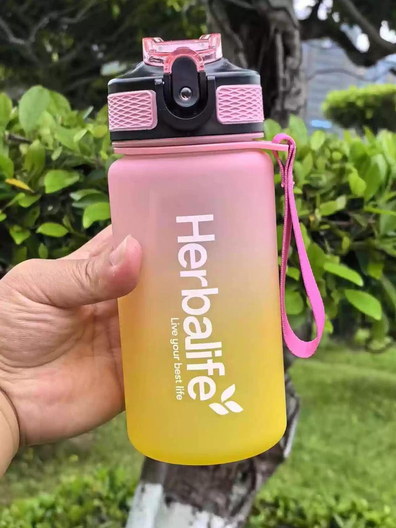 May include: A pink and yellow water bottle with a black lid and a pink carrying strap. The bottle has the text "Herbalife Live your best life" printed on it.