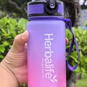 May include: A purple and pink water bottle with a black lid and a purple strap. The bottle has the Herbalife logo and the text "Live your best life".