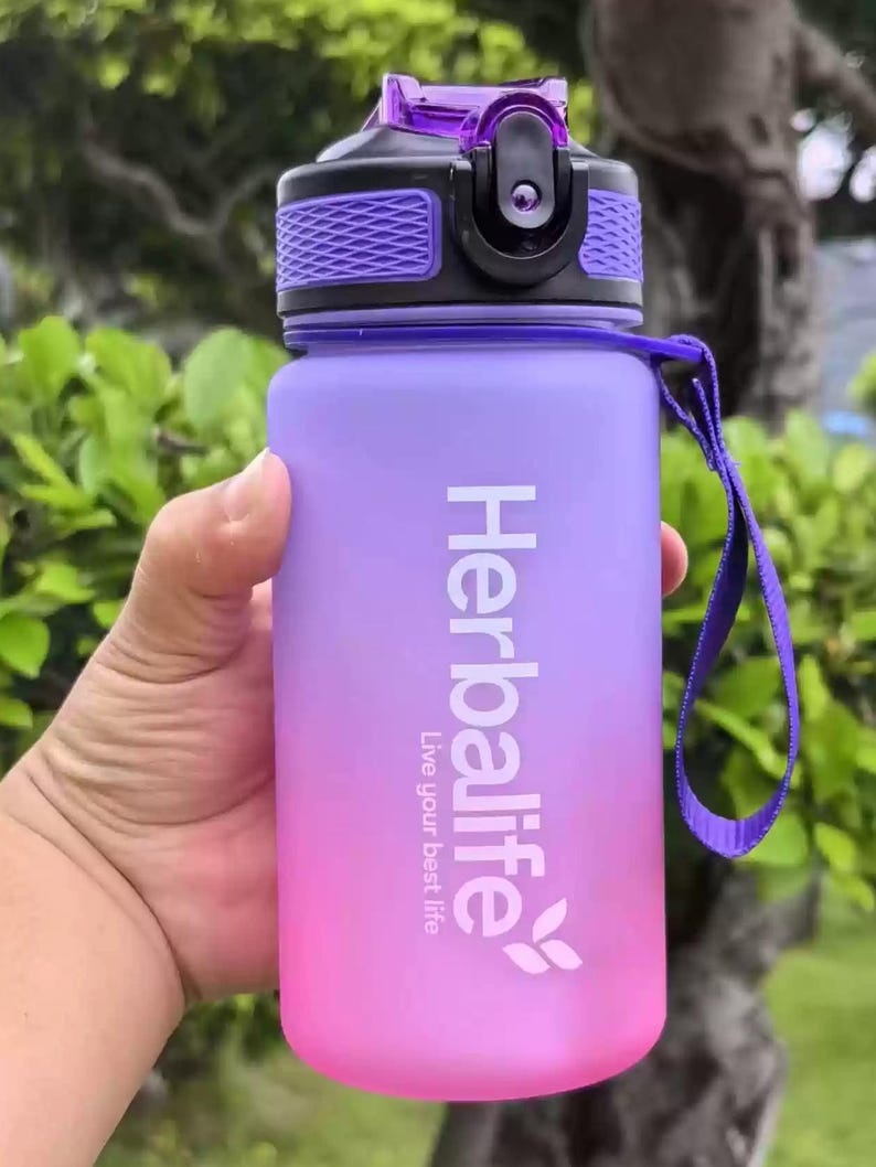 May include: A purple and pink water bottle with a black lid and a purple strap. The bottle has the Herbalife logo and the text "Live your best life".