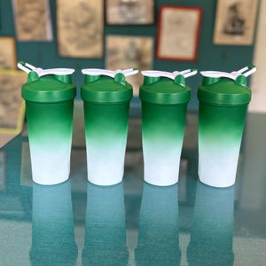 Puede incluir: Cuatro botellas de coctelera con degradado verde y blanco con tapas blancas y cierres abatibles. Las botellas son cil&iacute;ndricas y parecen ser de pl&aacute;stico. El fondo est&aacute; borroso.