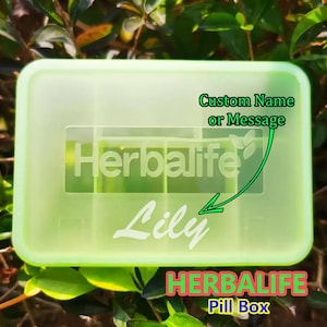 May include: A light green rectangular pill box with the Herbalife logo and the name "Lily" in white. The text "Custom Name or Message" is also visible, along with "HERBALIFE Pill Box". The box is surrounded by green foliage.