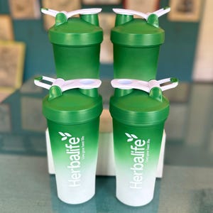 Puede incluir: Cuatro botellas de coctelera Herbalife verdes y blancas con tapas blancas y cierres abatibles. Las botellas tienen un dise&ntilde;o degradado, que pasa de verde a blanco. El logotipo y el texto de Herbalife est&aacute;n impresos en la parte delantera.