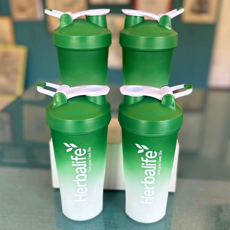 Puede incluir: Cuatro botellas de coctelera Herbalife verdes y blancas con tapas blancas y cierres abatibles. Las botellas tienen un dise&ntilde;o degradado, que pasa de verde a blanco. El logotipo y el texto de Herbalife est&aacute;n impresos en la parte delantera.