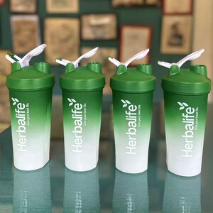 Puede incluir: Cuatro cocteleras Herbalife verdes y blancas con tapas y agitadores blancos. Las cocteleras tienen el logotipo de Herbalife y el texto "Live your best life". Las cocteleras est&aacute;n dispuestas en fila sobre una superficie reflectante.
