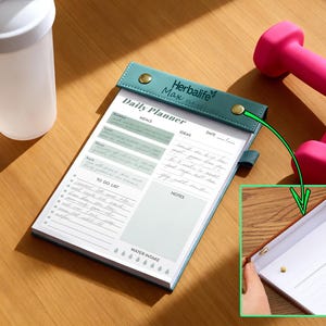 K&ouml;nnte beinhalten: Ein Tagesplaner mit tealfarbenem Einband und goldenen Knopfverschl&uuml;ssen, mit Abschnitten f&uuml;r Mahlzeiten, Ideen und eine To-Do-Liste. Eine wei&szlig;e Wasserflasche und rosa Hanteln sind ebenfalls im Bild. Der Planer tr&auml;gt den Text "Herbalife Max" und "Daily Planner".