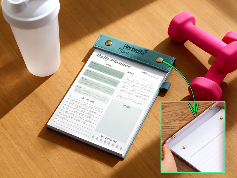 K&ouml;nnte beinhalten: Ein Tagesplaner mit tealfarbenem Einband und goldenen Knopfverschl&uuml;ssen, mit Abschnitten f&uuml;r Mahlzeiten, Ideen und eine To-Do-Liste. Eine wei&szlig;e Wasserflasche und rosa Hanteln sind ebenfalls im Bild. Der Planer tr&auml;gt den Text "Herbalife Max" und "Daily Planner".