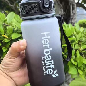 May include: A black and gray water bottle with a black lid and a black strap. The bottle has a white gradient and the text "Herbalife Live your best life" printed on it.