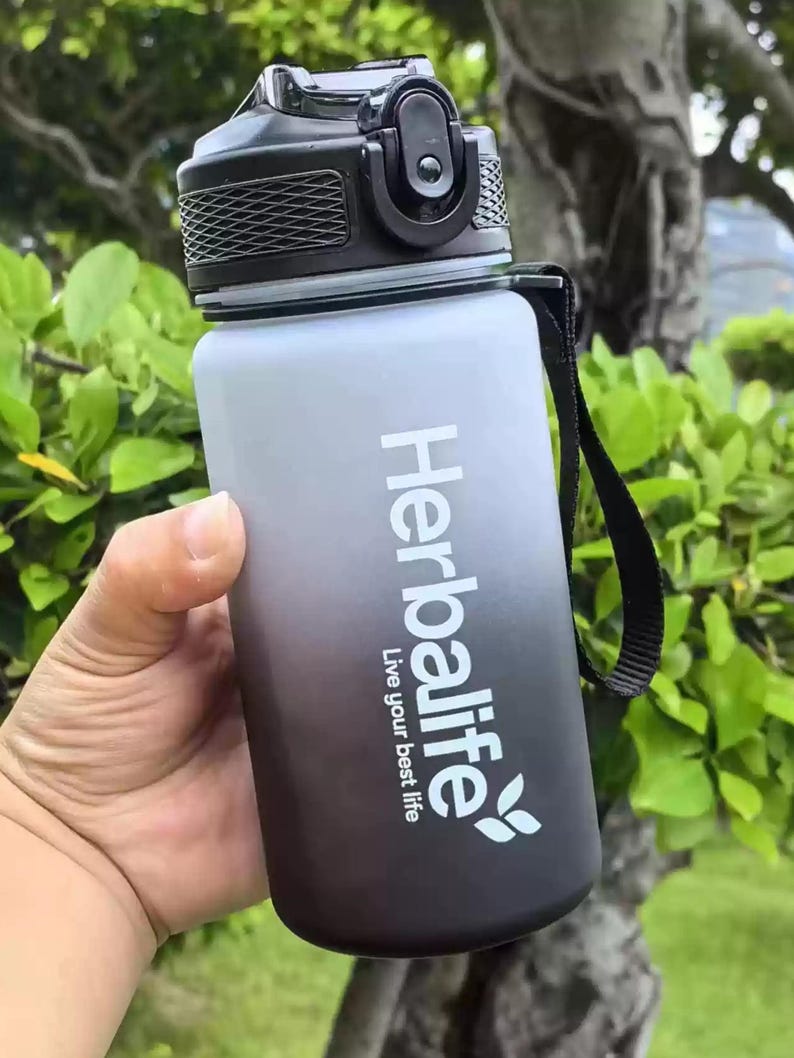May include: A black and gray water bottle with a black lid and a black strap. The bottle has a white gradient and the text "Herbalife Live your best life" printed on it.