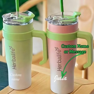 Könnte beinhalten: Zwei grüne und weiße Reisebecher mit grünem Griff und Deckel. Die Becher haben ein Farbverlaufsdesign mit den Worten "Herbalife" und "Benutzerdefinierter Name" auf dem linken Becher und "Herbalife" und "Julia" auf dem rechten Becher.