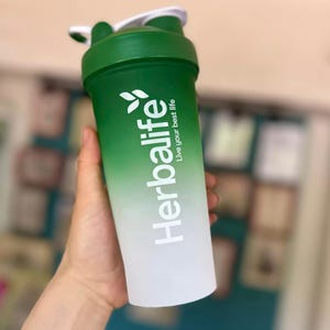 Puede incluir: Una botella mezcladora Herbalife verde y blanca. La botella tiene un dise&ntilde;o degradado, que pasa de verde en la parte superior a blanco en la inferior. El logotipo de Herbalife y el texto "Live your best life" est&aacute;n impresos en blanco.