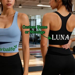 May include: A light blue sports bra with a navy blue and white band featuring the text "Herbalife" in green. The model is wearing black leggings. The back of the bra is black with the name "LUNA" in white.