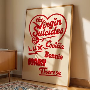 May include: A framed poster with the text "The Virgin Suicides" in red, set against a cream background. The names Lux, Cecilia, Bonnie, Mary, and Therese are also displayed. The poster leans against a wall near a wooden cabinet and a patterned rug.