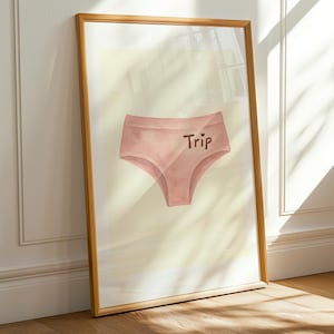 May include: A framed print featuring a watercolor illustration of pink underwear with the word "Trip" printed on the front. The artwork is displayed against a light yellow background, leaning against a white wall and wooden floor.