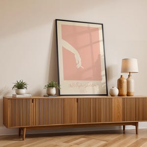 Peut inclure: Un meuble TV en bois avec un design &agrave; lattes et deux portes. Le meuble est dans un salon avec un poster encadr&eacute; repr&eacute;sentant un fond rose et une silhouette de main tenant une cigarette. Il y a deux plantes en pot et une lampe sur le meuble.