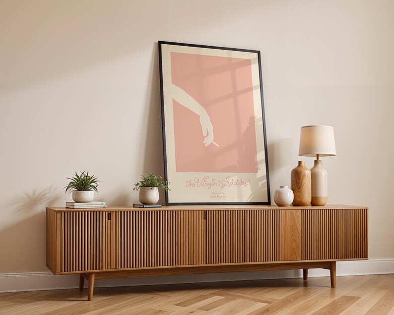 Peut inclure: Un meuble TV en bois avec un design &agrave; lattes et deux portes. Le meuble est dans un salon avec un poster encadr&eacute; repr&eacute;sentant un fond rose et une silhouette de main tenant une cigarette. Il y a deux plantes en pot et une lampe sur le meuble.