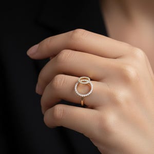 May include: A gold ring with two overlapping circles, each studded with small, clear gemstones. The ring is shown against a dark backdrop, accentuating its refined design. The ring's band is a slender, polished gold.