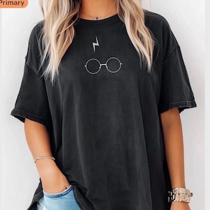 Puede incluir: Camiseta negra con un rayo blanco y gafas redondas. La camiseta de manga corta tiene un corte holgado y un aspecto ligeramente descolorido. El gráfico está centrado en el pecho.