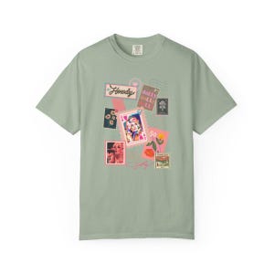 K&ouml;nnte beinhalten: Salbeigr&uuml;nes T-Shirt mit einer Collage von Grafiken im Briefmarkenstil. Das Design enth&auml;lt das Wort "Howdy", ein Portr&auml;t und den Namen "Dolly" in Schriftform. Das Shirt hat einen klassischen Rundhalsausschnitt und kurze &Auml;rmel.