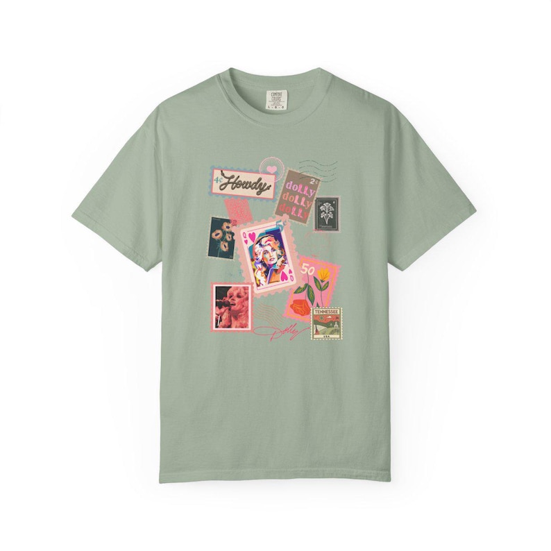 K&ouml;nnte beinhalten: Salbeigr&uuml;nes T-Shirt mit einer Collage von Grafiken im Briefmarkenstil. Das Design enth&auml;lt das Wort "Howdy", ein Portr&auml;t und den Namen "Dolly" in Schriftform. Das Shirt hat einen klassischen Rundhalsausschnitt und kurze &Auml;rmel.