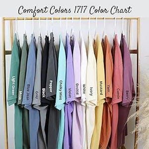K&ouml;nnte beinhalten: Eine Farbkarte zeigt eine Auswahl an T-Shirts, die auf einem Gestell h&auml;ngen. Die Shirts sind in verschiedenen Farben erh&auml;ltlich, darunter Hellgr&uuml;n, Blue Jean, Schwarz, Violett und Senf. Der Text "Comfort Colors 1717 Color Chart" steht oben.