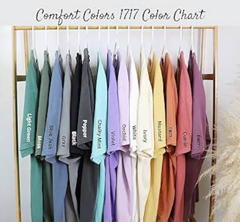 K&ouml;nnte beinhalten: Eine Farbkarte zeigt eine Auswahl an T-Shirts, die auf einem Gestell h&auml;ngen. Die Shirts sind in verschiedenen Farben erh&auml;ltlich, darunter Hellgr&uuml;n, Blue Jean, Schwarz, Violett und Senf. Der Text "Comfort Colors 1717 Color Chart" steht oben.