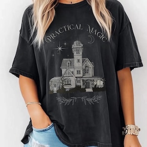 Puede incluir: Camiseta negra con las palabras "PRACTICAL MAGIC" sobre una ilustración detallada de una casa con una torre, estrellas y una luna creciente. El diseño es de estilo vintage.