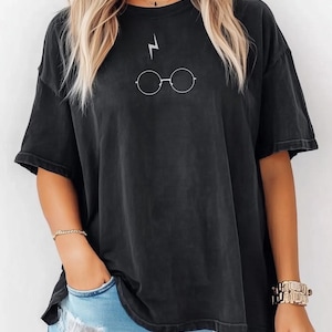 May include: Black t-shirt with a white lightning bolt and round glasses design. The top has short sleeves and a loose fit. The model is wearing blue jeans.