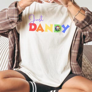 Puede incluir: Camiseta color crema con el texto "Just Dandy" en letras coloridas y un gráfico de flores. La camiseta se combina con pantalones cortos negros, calcetines a rayas y una camisa a cuadros marrón.