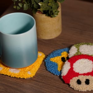 May include: Four crocheted coasters featuring different video game characters. The coasters are yellow, blue, green, and red. A blue mug is sitting on the yellow coaster.