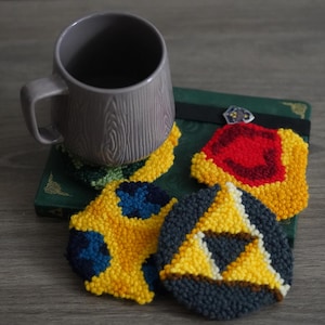 May include: Four crocheted coasters featuring different designs from the Legend of Zelda video game series. The coasters are yellow, blue, red, and black with white accents. A gray mug sits on top of one of the coasters.