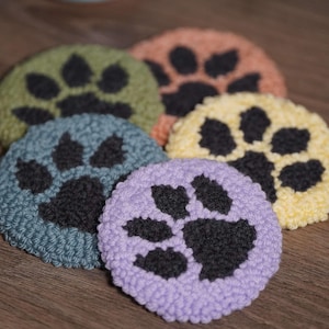 May include: Five crocheted coasters with paw prints in black. The coasters are in various colors, including green, blue, purple, yellow, and orange.