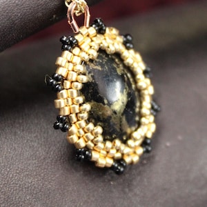 May include: A gold-toned necklace with an oval pendant. The pendant features a dark, speckled stone surrounded by gold and black beads. The necklace is resting on a dark brown surface.