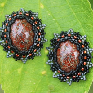 May include: Oval-shaped stud earrings with a red and brown stone center, surrounded by black, orange, and gray beads. The earrings are set against a green leaf background.