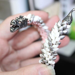 Op de afbeelding: Een zilveren drakenarmband met een gedetailleerde drakenkop en een haaksluiting. De armband is gemaakt van witte, zilveren en roze kralen, wat een uniek ontwerp cre&euml;ert. De ogen van de draak zijn rood.