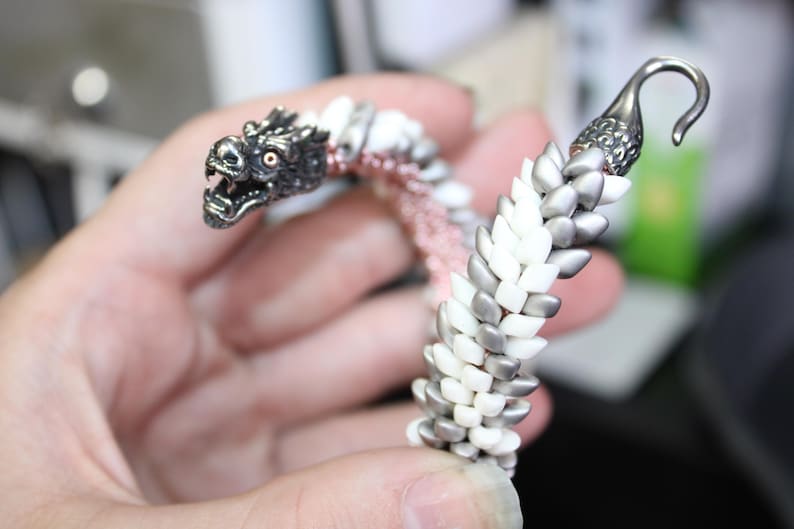 Op de afbeelding: Een zilveren drakenarmband met een gedetailleerde drakenkop en een haaksluiting. De armband is gemaakt van witte, zilveren en roze kralen, wat een uniek ontwerp cre&euml;ert. De ogen van de draak zijn rood.